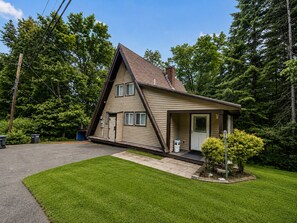Une maison de plain-pied avec un toit en pente, un porche à l’avant et un petit jardin devant, avec un arbre.