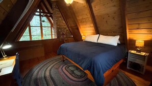 Um quarto de sótão acolhedor com teto inclinado, uma cama grande com colcha azul, uma escrivaninha de madeira com candeeiro e uma janela com vista para árvores.