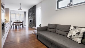 Uma sala de estar moderna com um sofá cinzento, piso de madeira, uma parede de destaque em pedra, uma área de jantar com mesa e cadeiras, e uma cozinha com armários brancos e uma janela.