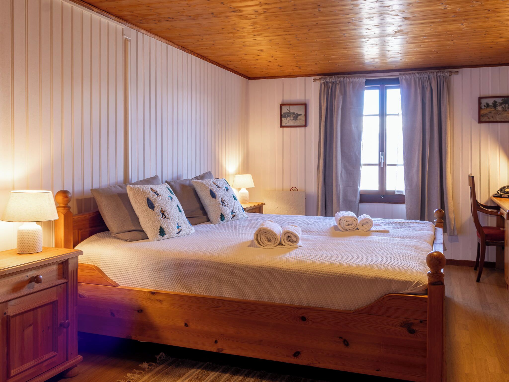 A neatly made bed with pillows, a wooden headboard, a nightstand with a lamp, a window with curtains, and a small table with a chair.