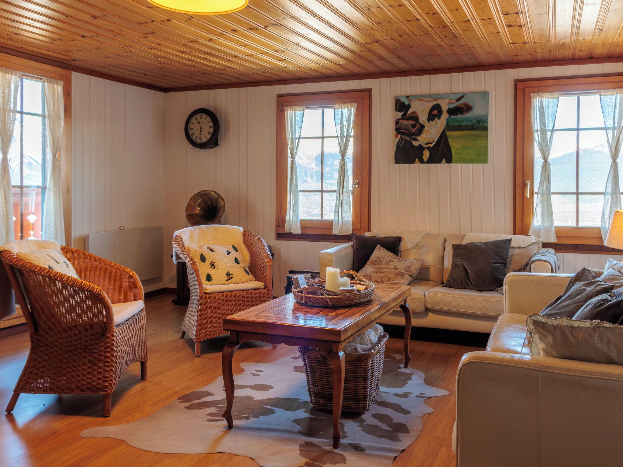 Un salon chaleureux avec un plafond en bois, un fauteuil en osier, un canapé, une table recouverte d’un tapis en peau de vache et un tableau représentant une vache.