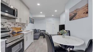 A modern kitchen with built-in appliances, a dining area with a white table and black chairs, and a living area with a sofa and a painting of a desert landscape. - Newly Built Guesthouse Suite (Calgary)