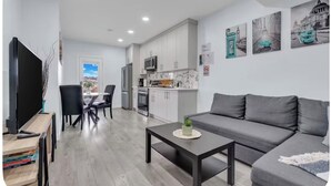 A modern living room with a grey sofa, a coffee table, and a TV. There is a kitchen area with white cabinets and a dining table with chairs. - Newly Built Guesthouse Suite (Calgary)