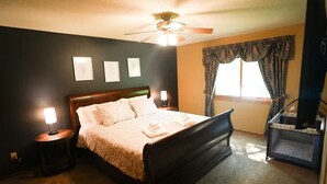 A bedroom with a bed, bedside lamps, a ceiling fan, a window with curtains, and a small table with a lamp.