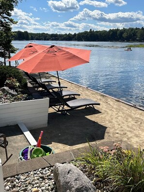 A lakeside patio with lounge chairs and a red umbrella, overlooking a calm lake. - Luxury Riverfront Sanctuary (Stanwood)