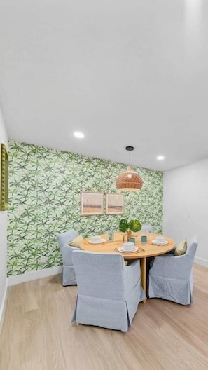 A dining area with a round table, two chairs, a hanging light fixture, and a wall with a leafy pattern.
