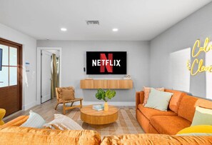 A modern living room with a flat-screen TV mounted on the wall, a cozy orange sofa, a wooden coffee table, and a wicker chair. - Wellness Retreat at WPB - HotTub | Sauna | ColdPlunge | Golf (West Palm Beach)