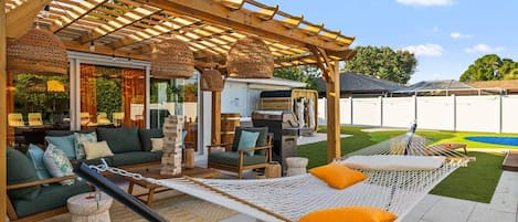 Un área de estar al aire libre techada con una parrilla, una hamaca y una piscina al fondo.