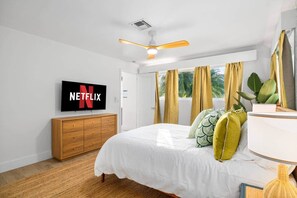 A bedroom with a bed, a wooden dresser, a television displaying the Netflix logo, a ceiling fan, and a window with curtains.
