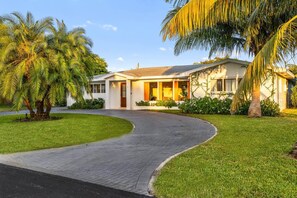 A single-story house with a curved driveway, surrounded by well-maintained grass and palm trees. - Wellness Retreat at WPB - HotTub | Sauna | ColdPlunge | Golf (West Palm Beach)