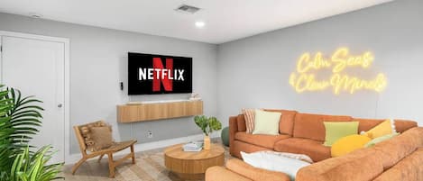 Un salon moderne avec un téléviseur connecté à Netflix, un néon, un canapé, une table basse et un fauteuil.