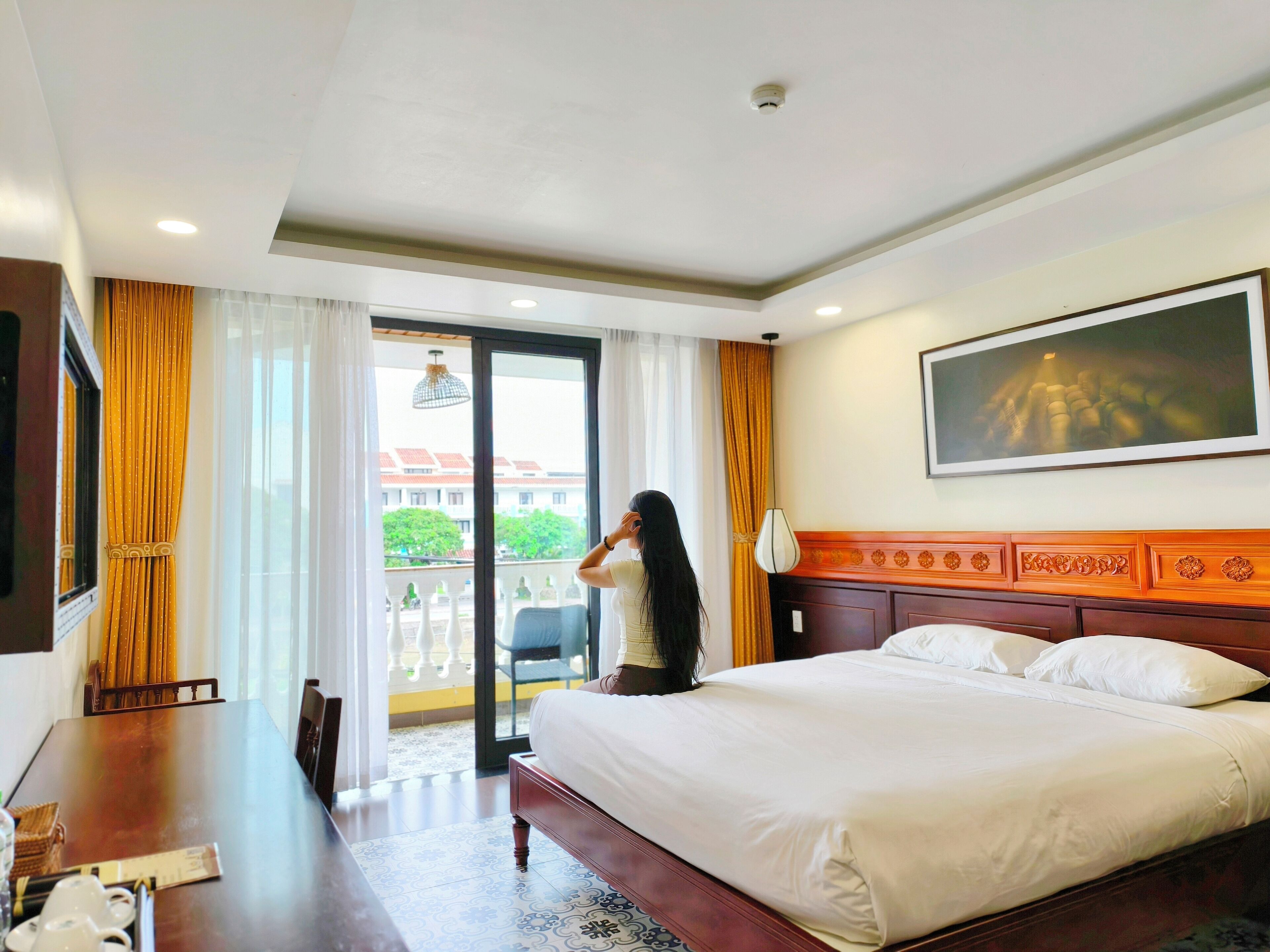 A hotel room with a large bed, a dining table, and a balcony with a view of buildings and trees.