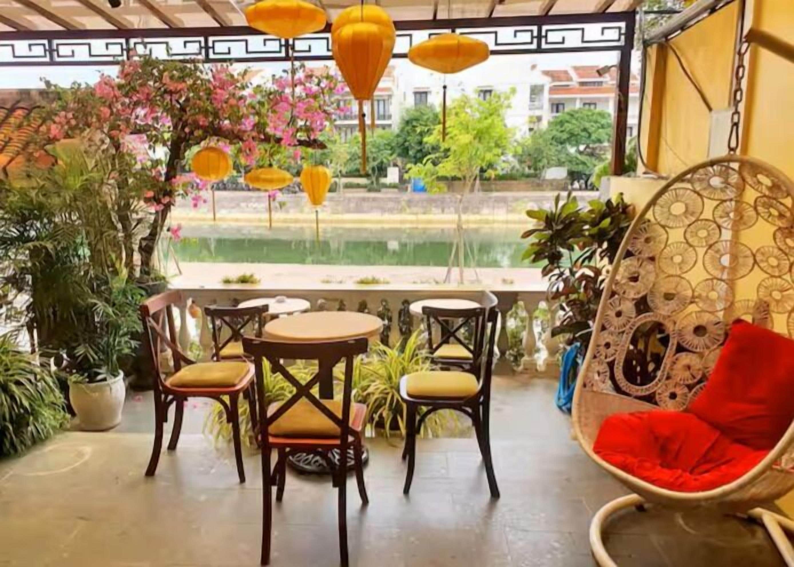 Un espacio al aire libre con mesas y sillas, un sillón colgante con cojín rojo, y vista a un cuerpo de agua y follaje.