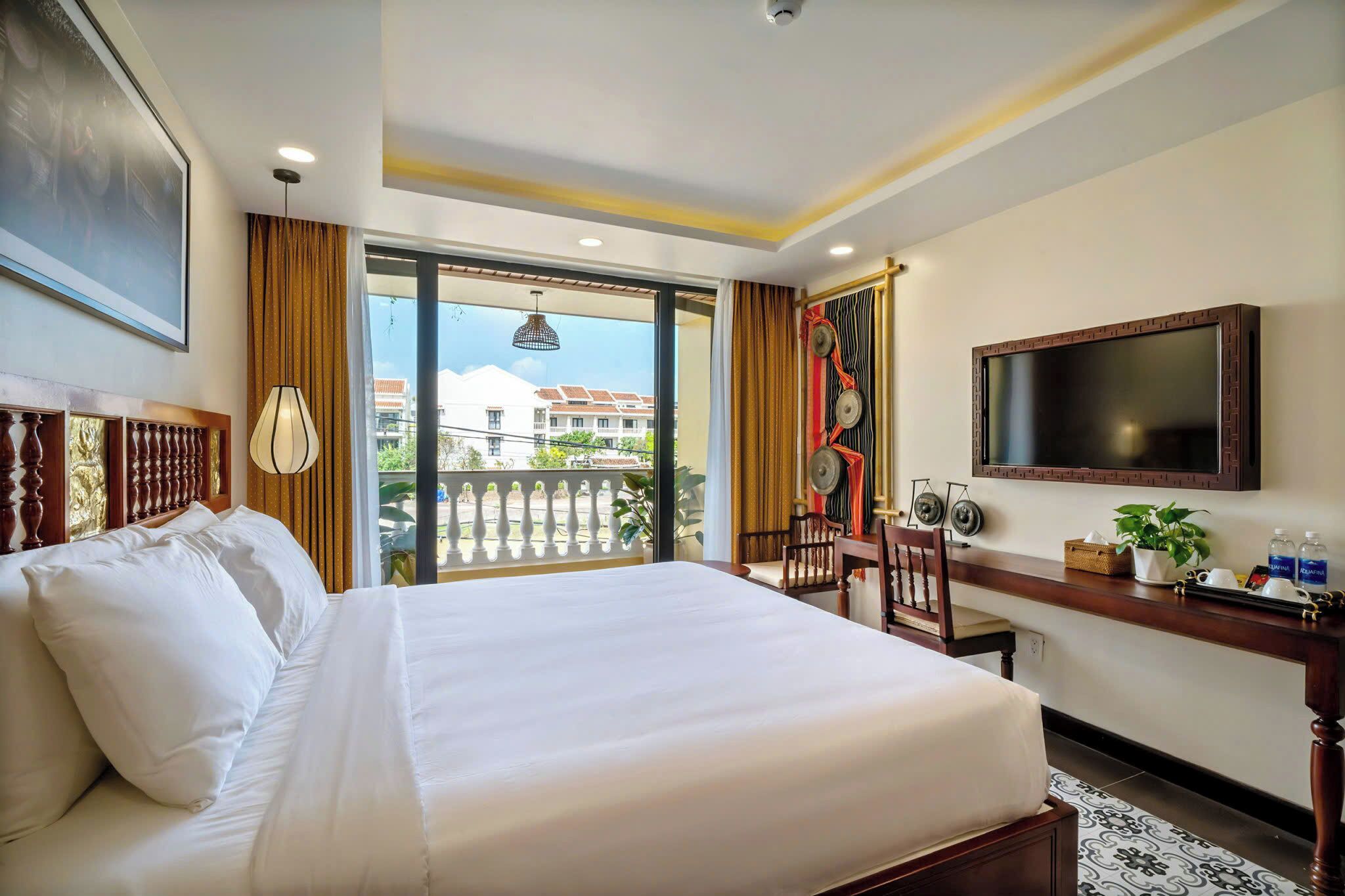 A hotel room with a large bed, a desk with a chair, a TV, and a balcony with a view of buildings.