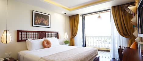 A hotel room with a large bed, a wooden headboard, a painting on the wall, a desk with a lamp, and a view of the outside through a window with curtains.