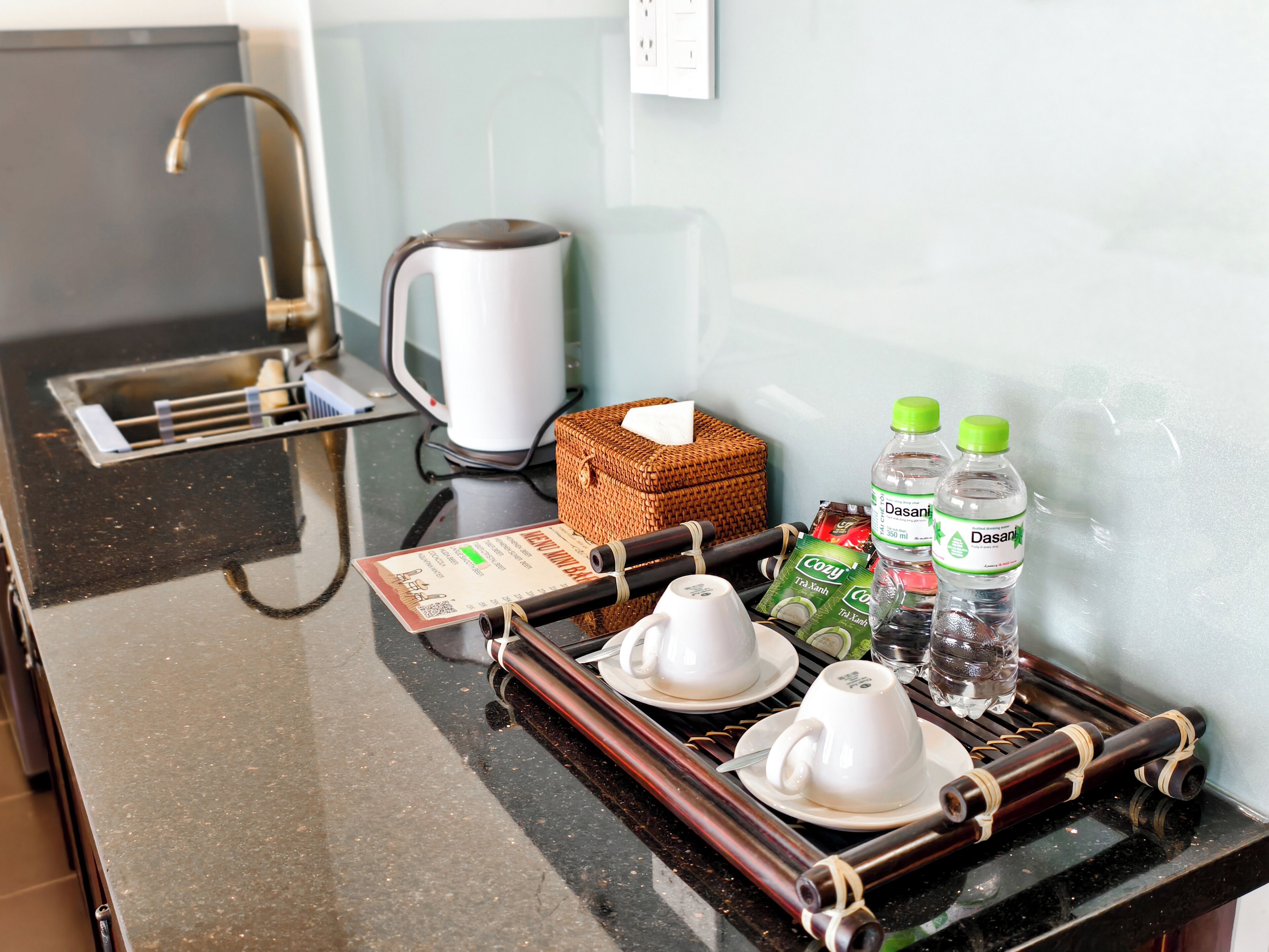 Bandeja de desayuno con tazas, una tetera y botellas de agua Dasani sobre una encimera de hotel.