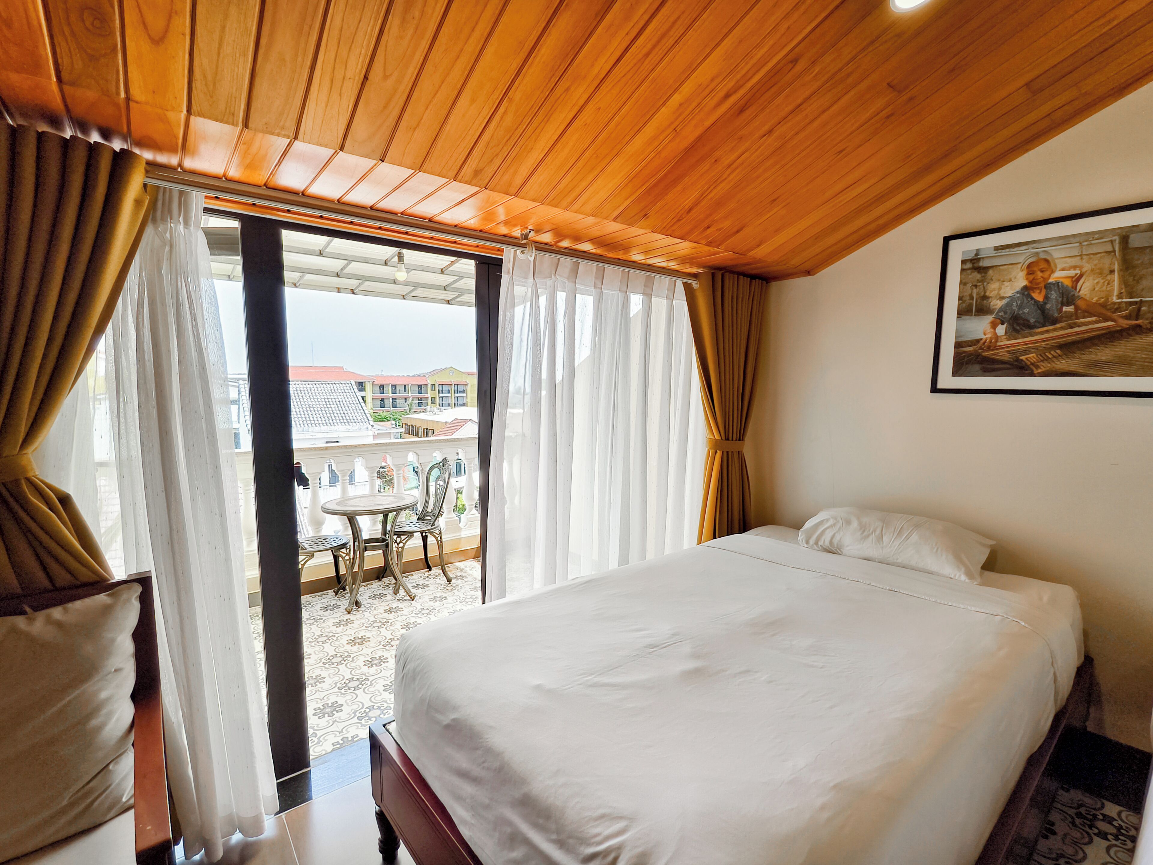 A hotel room with a bed, a chair, a table, and a balcony with a view of buildings.
