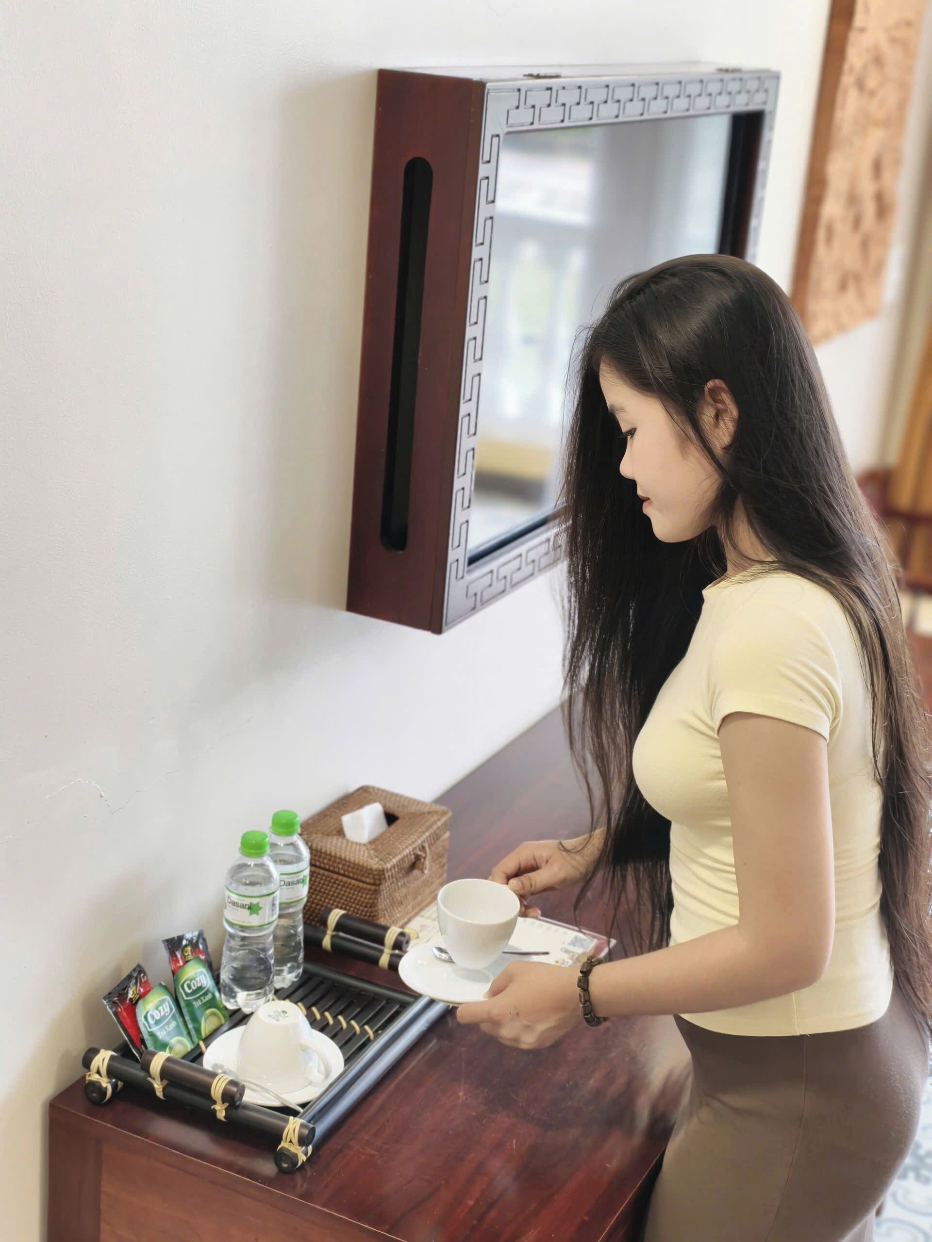 Una mujer con top amarillo y falda marrón está de pie junto a una mesa de madera con una bandeja que contiene una taza, una caja de pañuelos y agua embotellada.