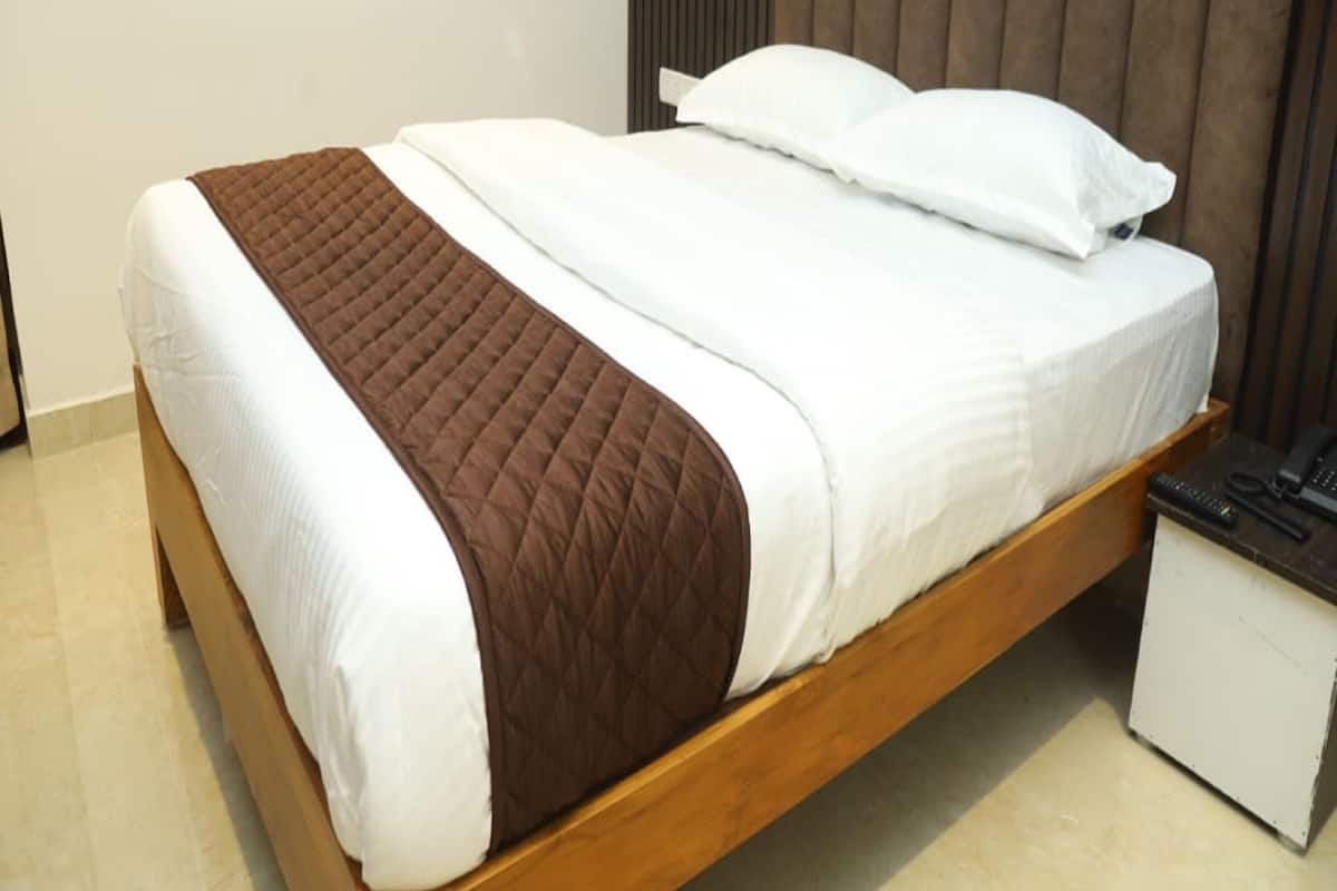 A neatly made bed with a brown quilted blanket and white sheets, a wooden headboard, and a small bedside table with a phone and remote controls.