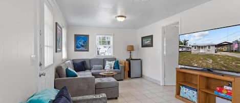 A living room with a grey sofa, a television, and a small table. There are windows and a door in the room.