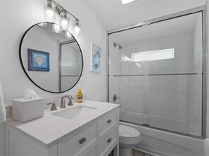 A modern bathroom with a white vanity, a round mirror with lights, a glass shower enclosure, and a toilet. - The Pelican Pad - Steinhatchee (Steinhatchee)