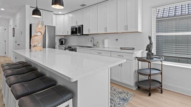A modern kitchen with white cabinets, a central island with black bar stools, pendant lighting, and a view of the outside through a window with blinds. - The Pelican Pad - Steinhatchee (Steinhatchee)