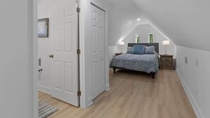 A bedroom with a sloped ceiling, a bed with a patterned bedspread, two bedside lamps, and a door with a picture frame. - The Pelican Pad - Steinhatchee (Steinhatchee)
