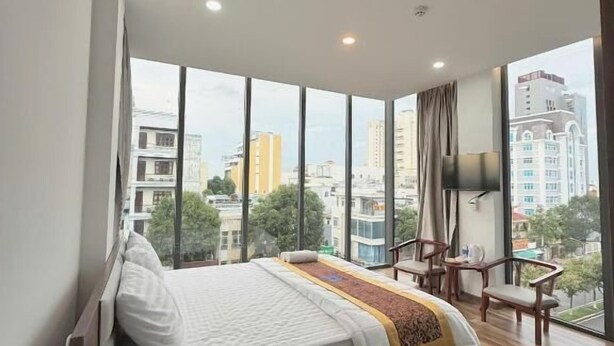 Una habitación de hotel moderna con una cama grande, un escritorio, una silla, ventanas amplias con vista a la ciudad y un televisor de pantalla plana montado en la pared.