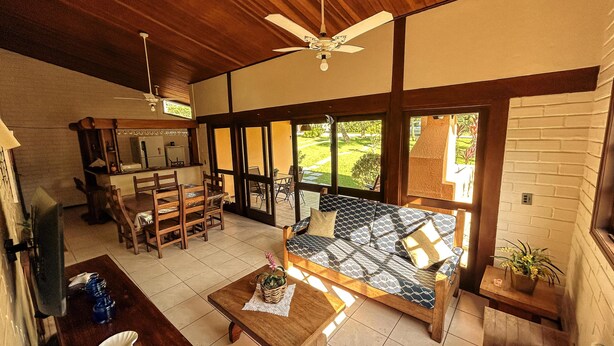 A spacious living area with a wooden ceiling, a ceiling fan, a sofa, a dining table, and a view of a garden through large windows. - House in the wonderful Pauba Condominium, a small village. (São Sebastião)