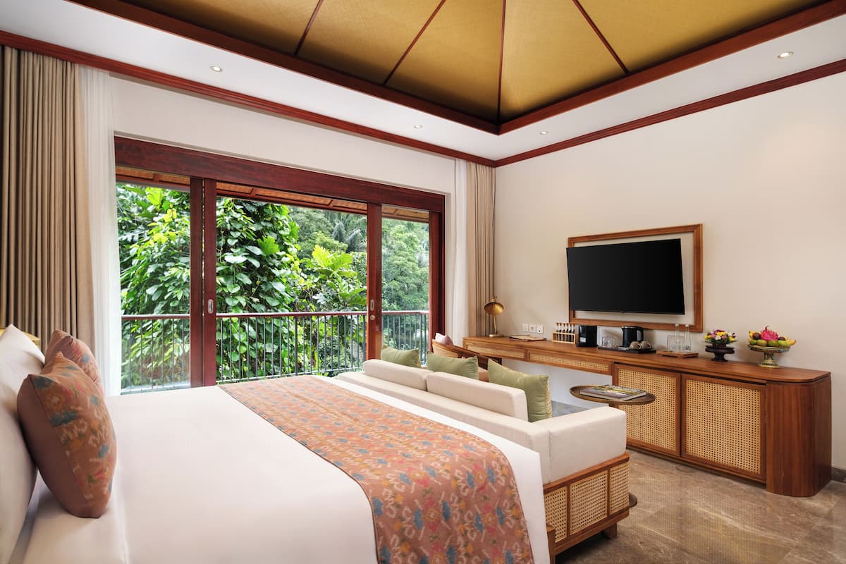 A bedroom with a large bed, a television, a desk with a computer, and a view of lush greenery.