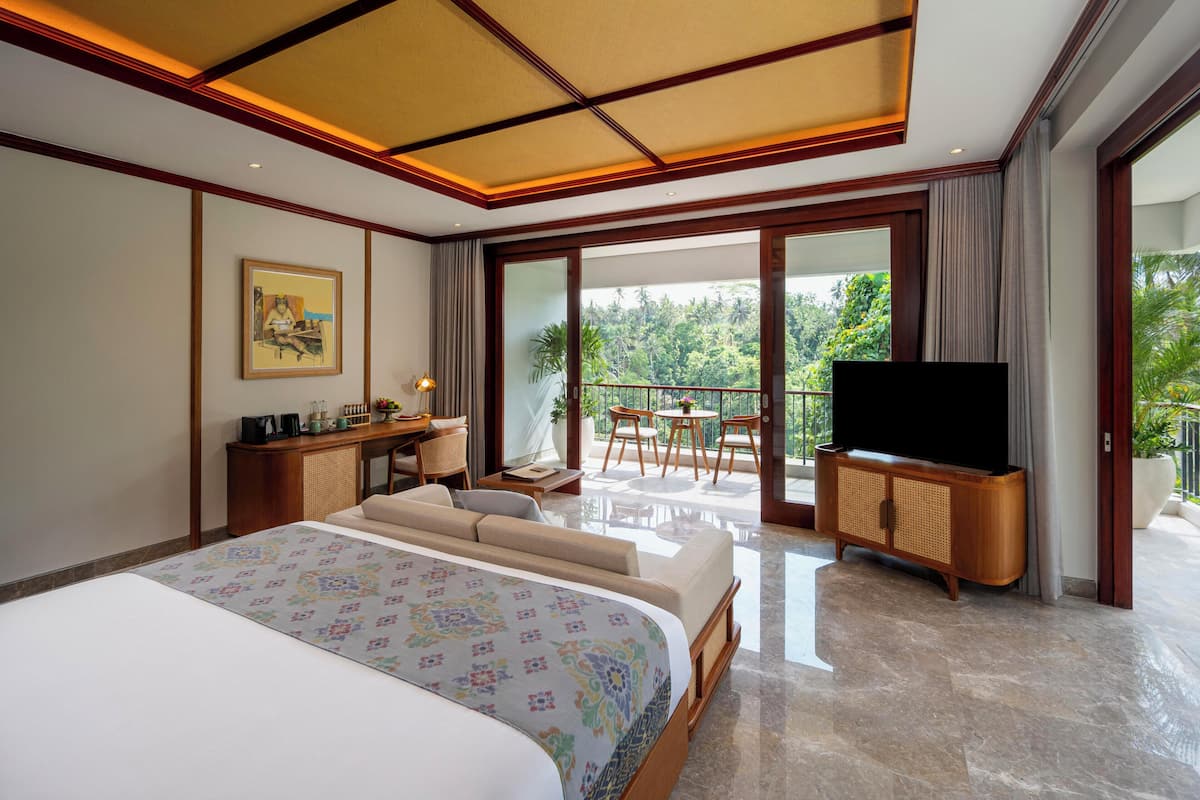 A hotel room with a bed, a television, a desk with a lamp, a chair, and a balcony with a view of greenery.