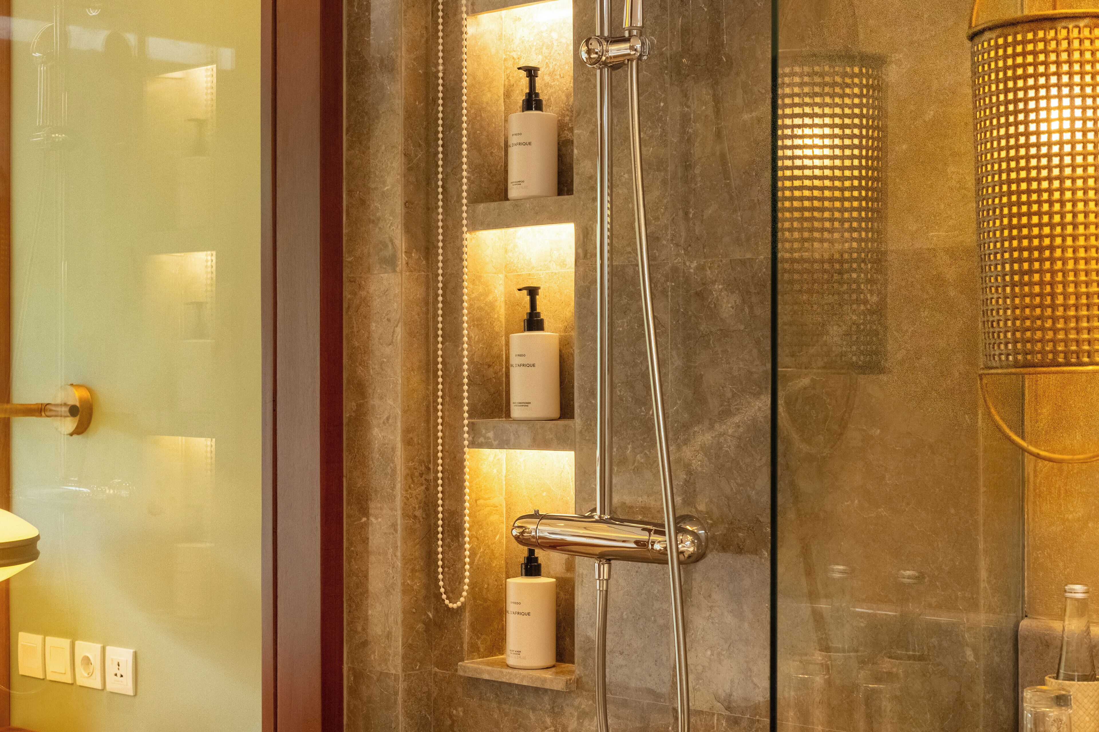 A bathroom with a glass door, a shower with a handheld showerhead, and shelves with toiletries.