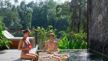A couple relaxing by a pool with a floating dining table, surrounded by lush greenery and a stone wall.