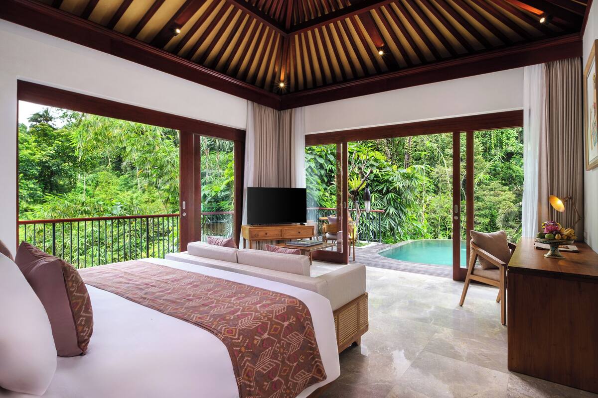 A bedroom with a large bed, a TV, a desk, and a view of a swimming pool and lush greenery.