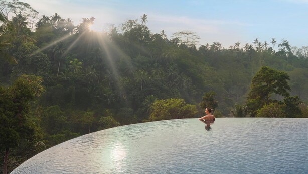 Una persona descansando en una amplia piscina curvada de borde infinito con vistas a exuberante follaje tropical y palmeras bajo un cielo despejado.