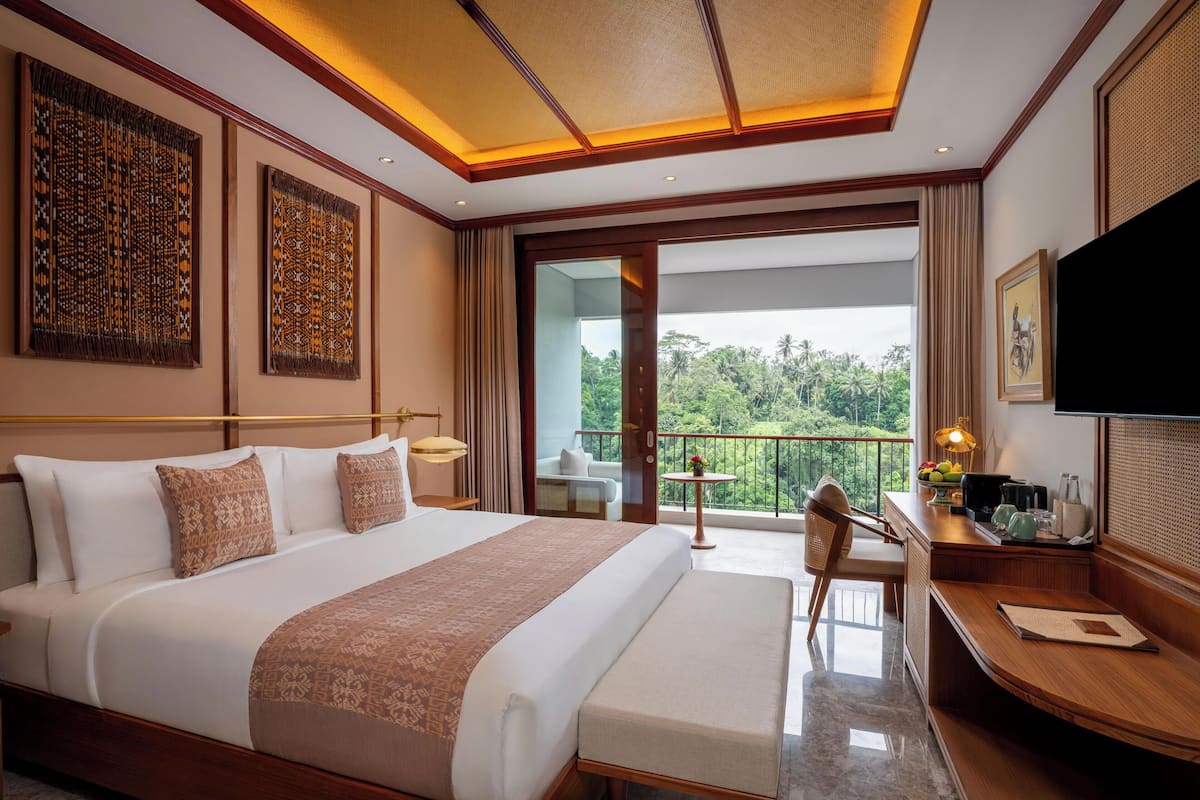 A hotel room with a large bed, a desk with a chair, a TV, and a balcony with a view of greenery.