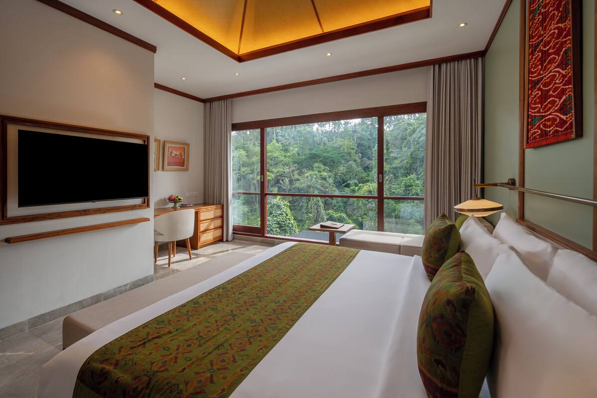 A hotel room with a large bed, a desk, a TV, and a window with a forest view.