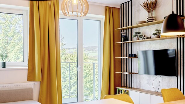 Un salon moderne avec un canapé, une table à manger, un téléviseur fixé au mur et une grande fenêtre avec des rideaux.