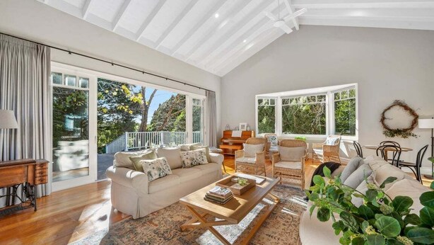 A spacious living room with a large sofa, a coffee table, and a dining area with chairs. There are large windows providing natural light and views of greenery outside.