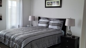 Ein Schlafzimmer mit einem Bett, zwei Nachttischen, einer Stehlampe, einer Deckenleuchte, einem Fenster mit Vorhängen und einem gerahmten Bild an der Wand.