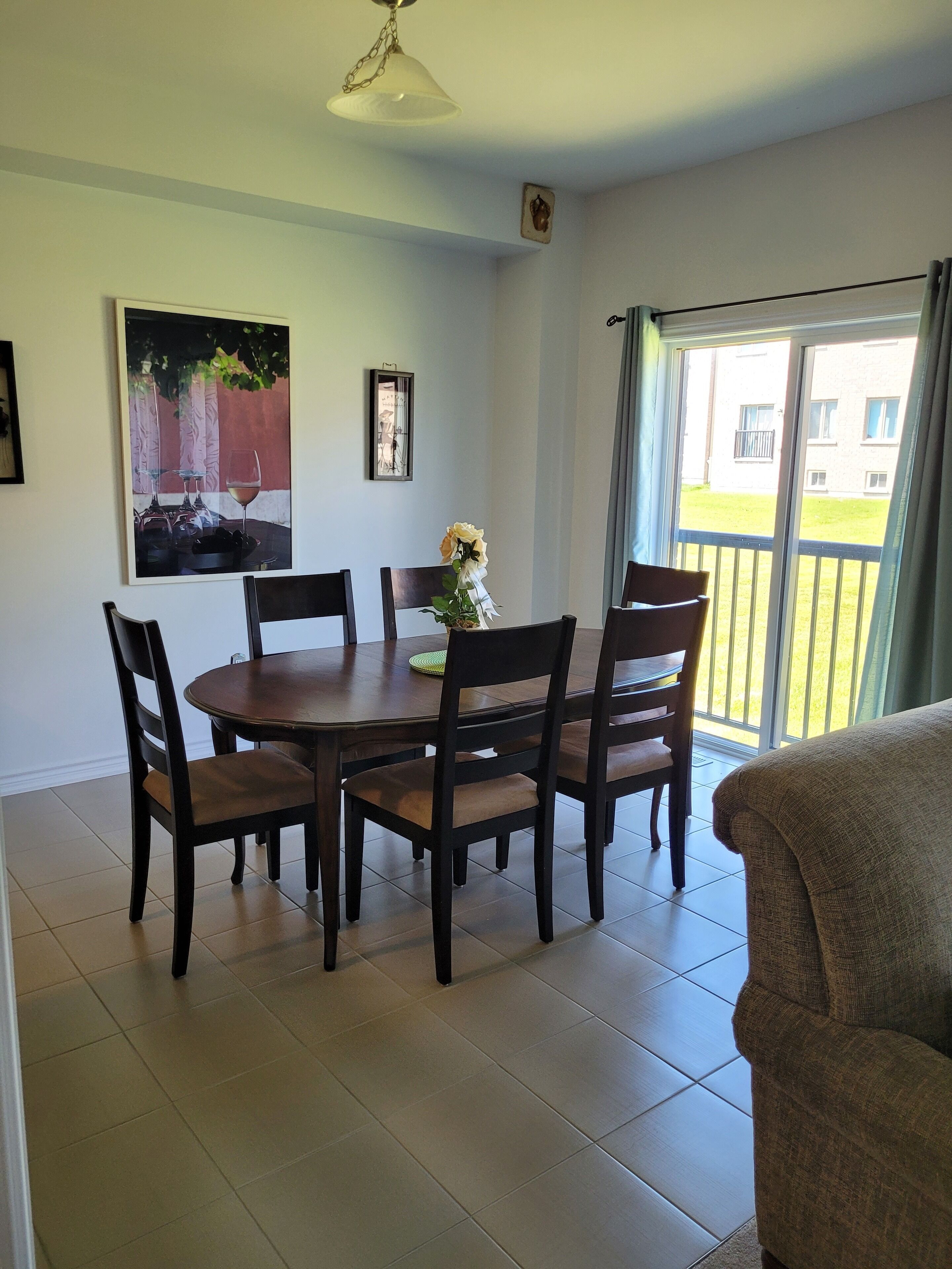 A dining area with a round table, six chairs, and a vase with flowers. There is a painting on the wall and a window with curtains.