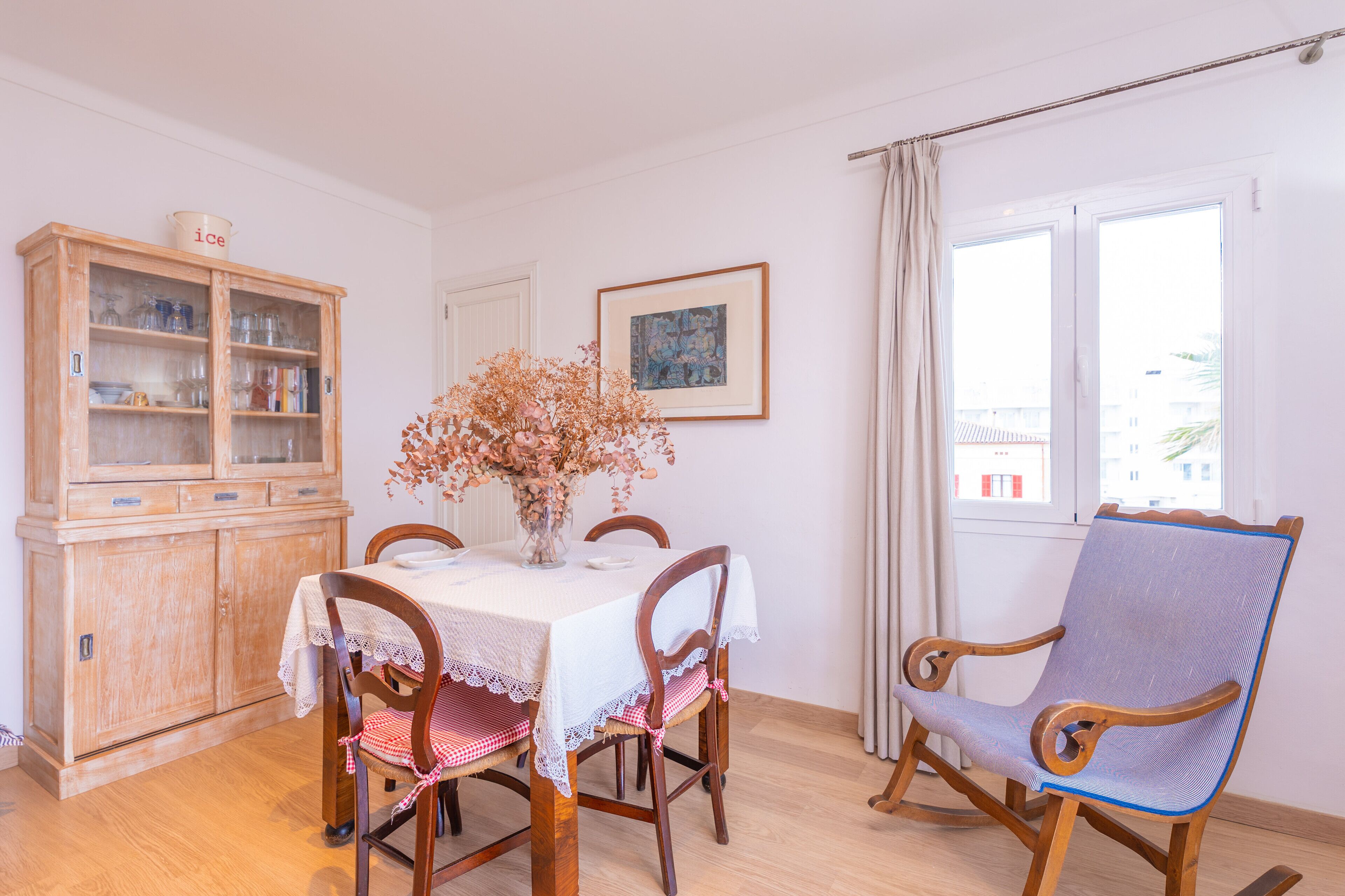 A dining area with a wooden cabinet, a table with a lace tablecloth, chairs, and a vase with dried flowers.