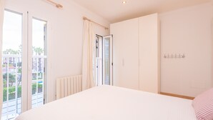 A bedroom with a large bed, a window with curtains, a radiator, and a view of buildings and greenery outside. - TUGORES SIMÓ 2º A - Charming apartment on the beachfront in Can Picafort – Free Wi-Fi (Muro)