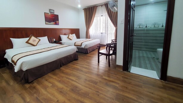 A hotel room with two beds, wooden flooring, a desk, and a sliding glass door leading to a balcony. - GOLD KIM PHÚ - Hotel Da Lat (Cam Ly - Da Lat)
