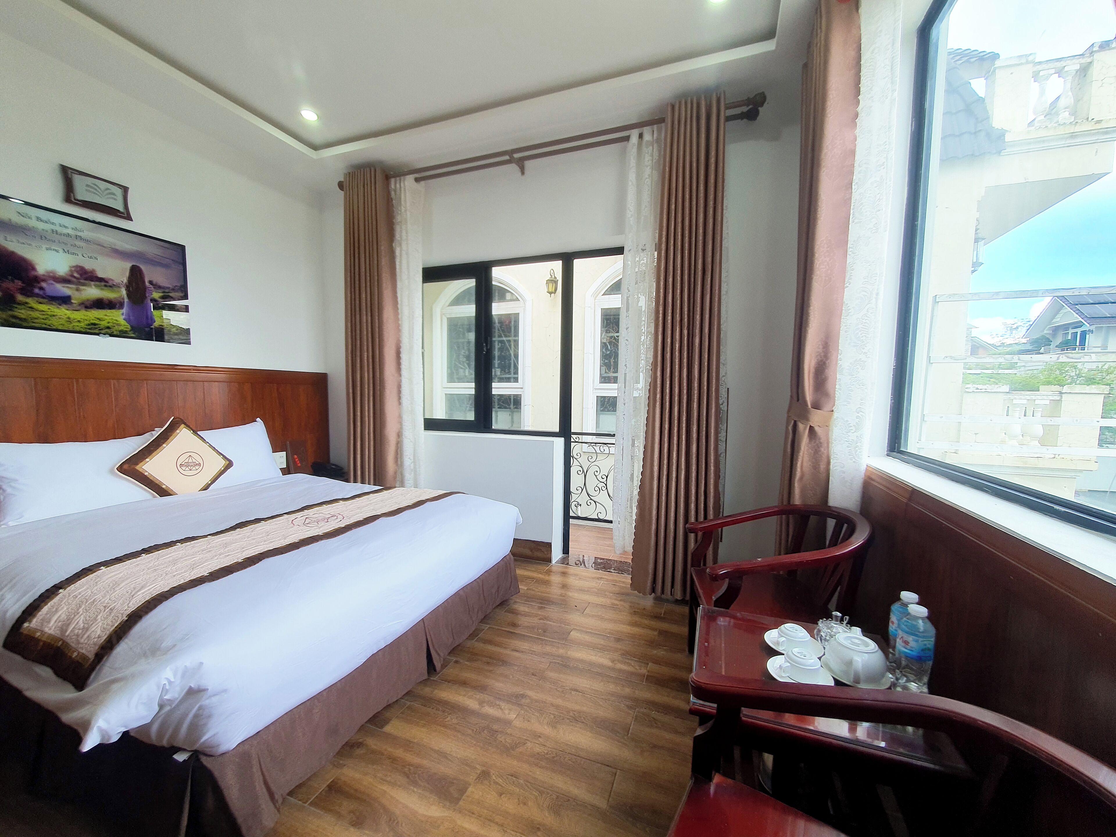 A hotel room with a bed, a wooden headboard, a window with curtains, a dining table with chairs, and a view of the outside.