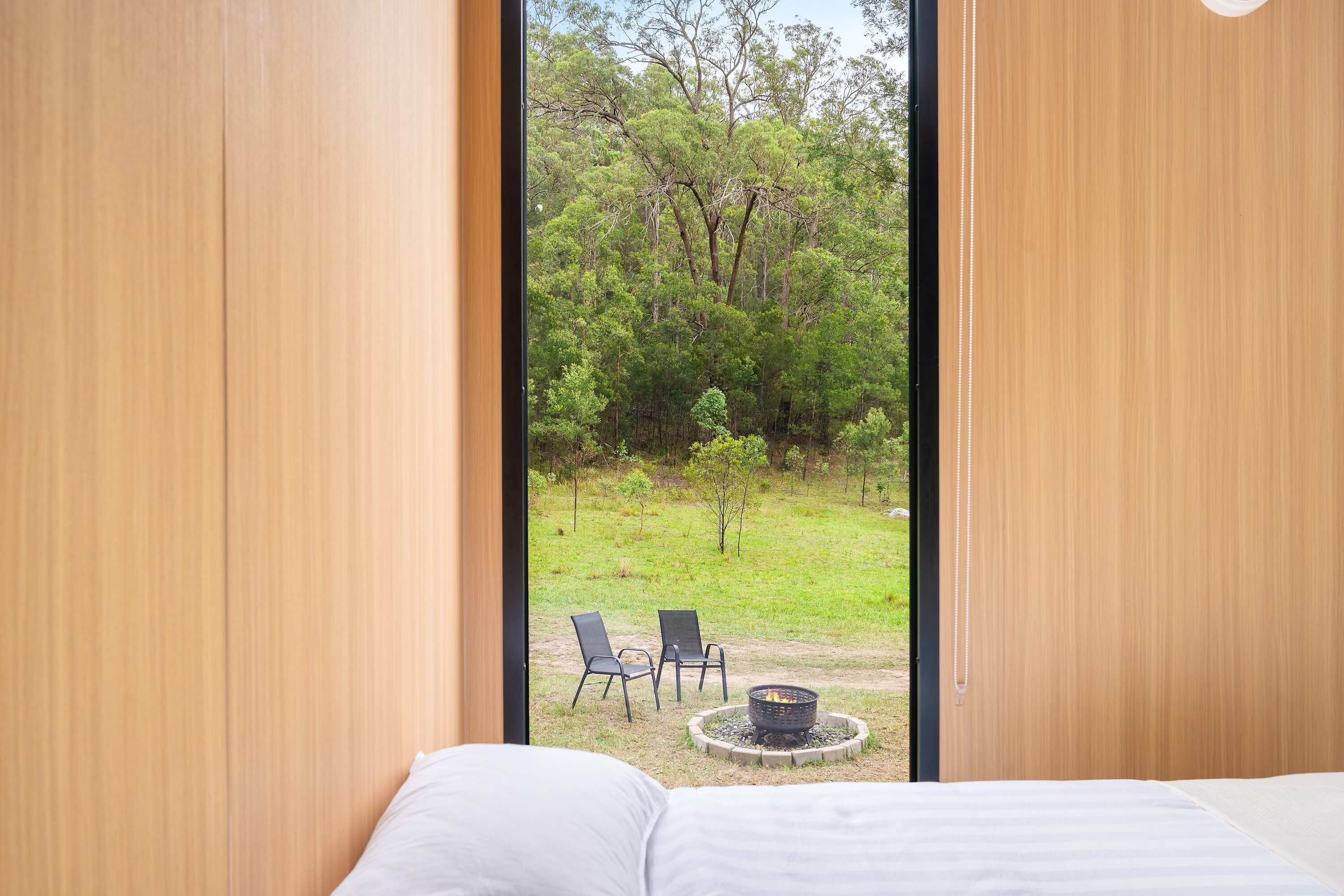 A bedroom with a large window, a bed, and a view of an outdoor area with chairs and a fire pit.