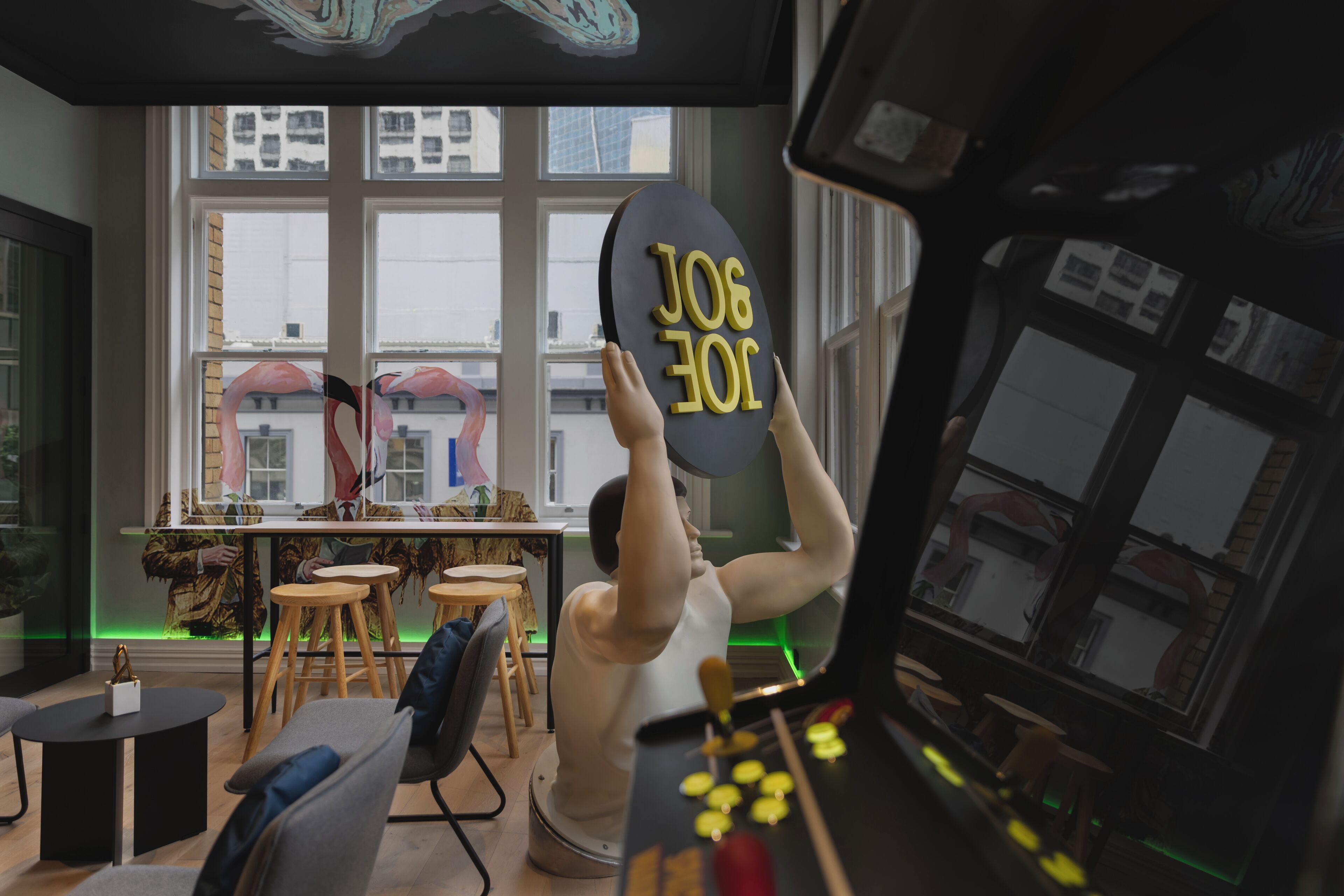Une personne tient une pancarte noire avec du texte jaune dans une pièce dotée d’une grande fenêtre, de tabourets en bois et d’une table.
