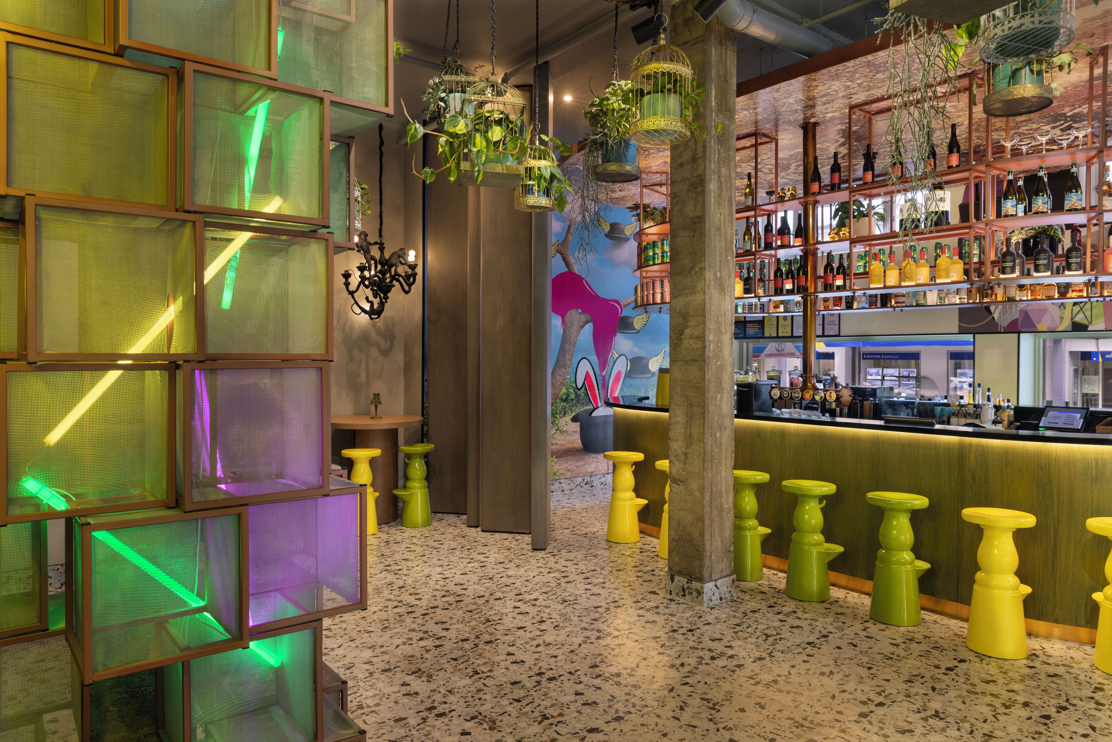 Zona de bar con piso moteado, jaulas colgantes con plantas y un mural de un árbol y pájaros.