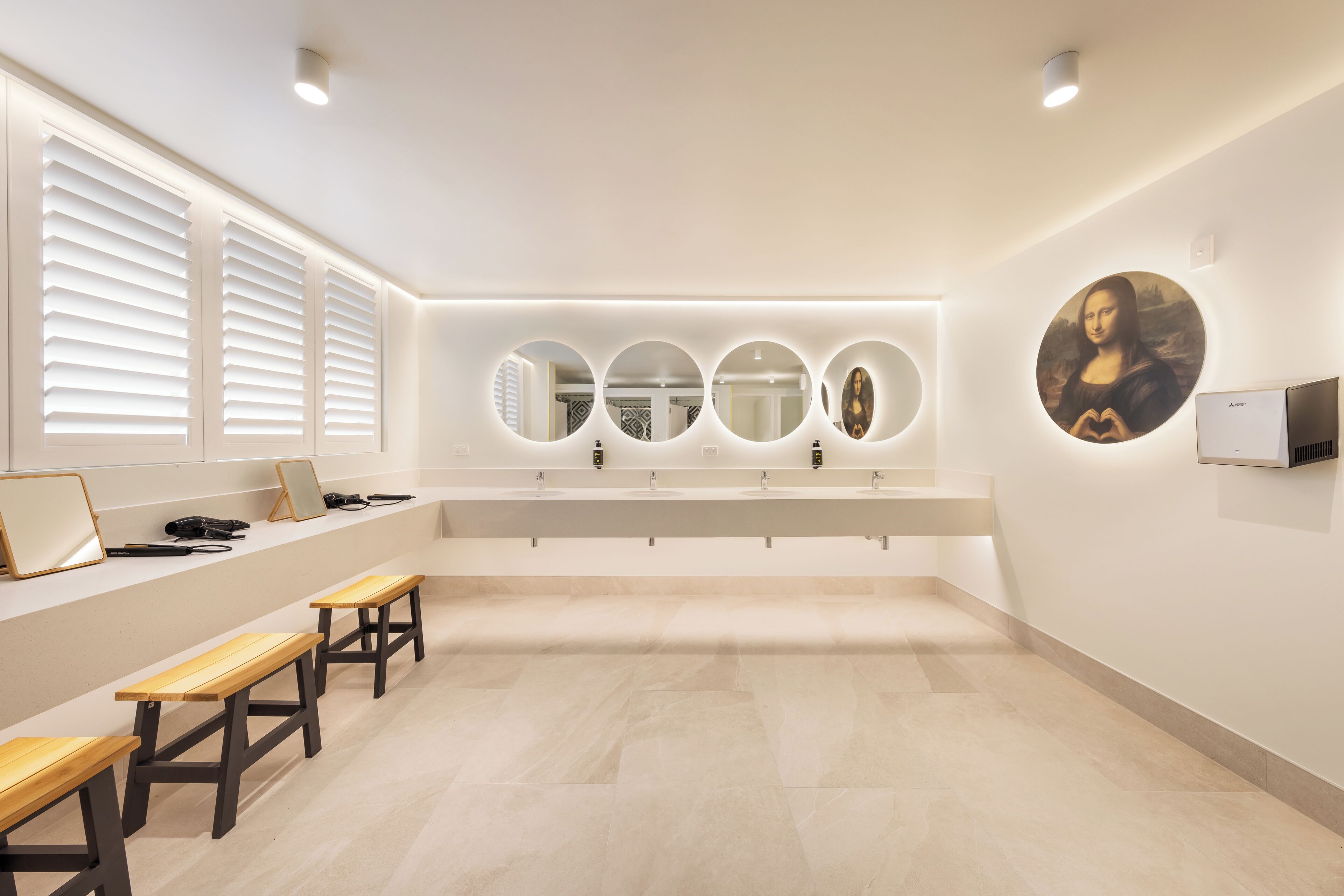 A modern bathroom with a large mirror, multiple sinks, wooden benches, and a wall-mounted artwork.