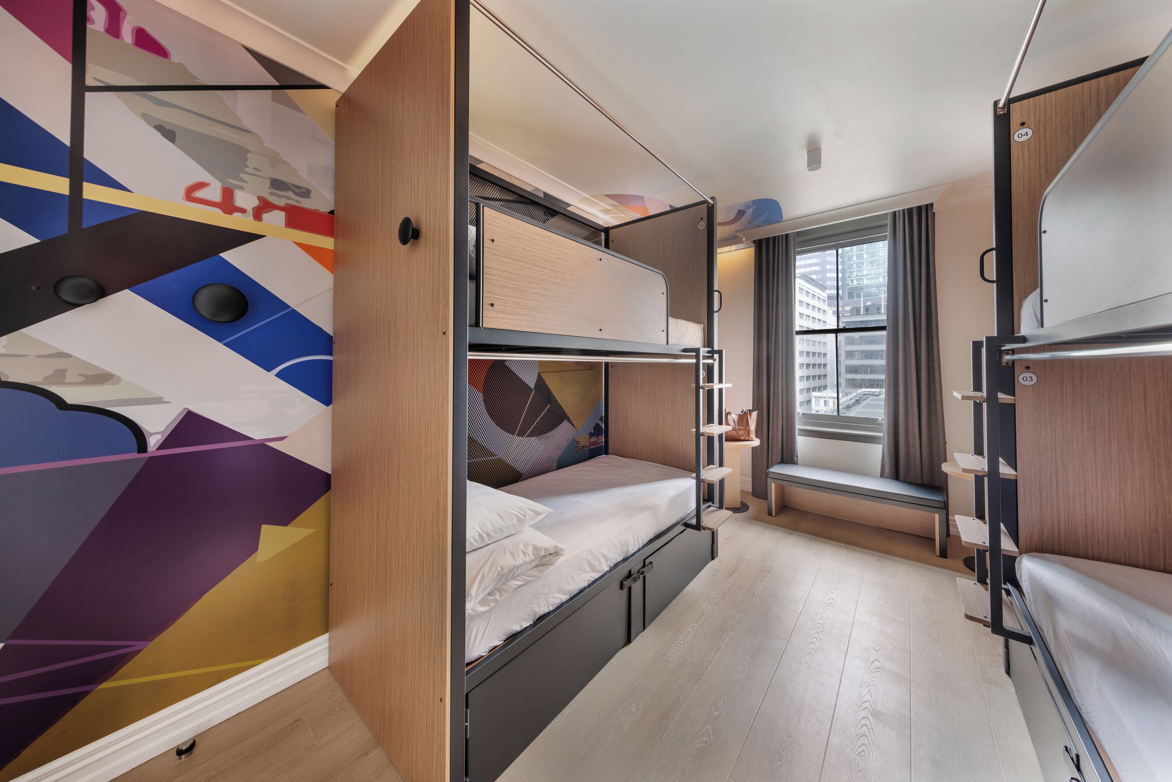 Una habitación compacta y moderna con una cama litera, un pequeño rincón de descanso junto a la ventana y un diseño geométrico y llamativo en la pared.